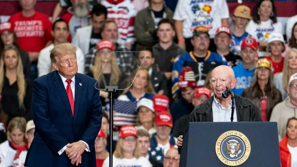 The image shows a public event where a man in a suit stands at a podium with the presidential seal, while another man in a suit listens attentively in front of a crowd of supporters.