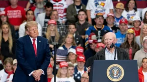 The image shows a public event where a man in a suit stands at a podium with the presidential seal, while another man in a suit listens attentively in front of a crowd of supporters.