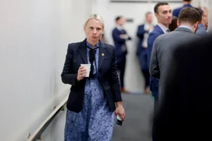 A woman in a professional outfit holds a cup and a phone as she walks through a hallway with other individuals in the background.