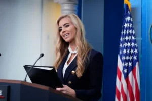 A woman in a professional setting, holding a folder, stands at a podium with an American flag in the background.