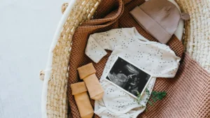 The image shows a baby's outfit, a beanie, and an ultrasound image arranged in a wicker basket, symbolizing pregnancy and preparation for a new life.