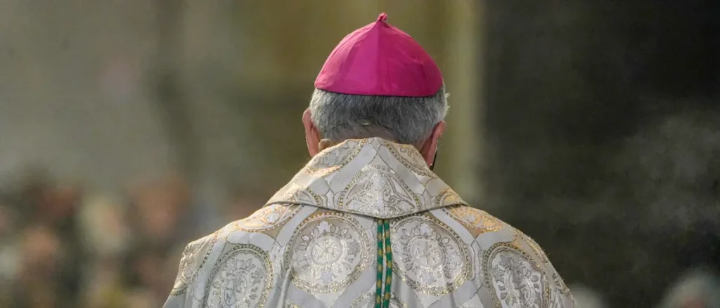 The image shows the back of a person wearing a white ornate vestment and a red mitre, likely a bishop, with a blurred crowd in the background.