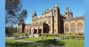 The image shows the Kelvingrove Art Gallery and Museum in Glasgow, Scotland, with people relaxing on the grass in front of the historic building on a sunny day.