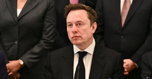 The image shows a man in a suit and tie, seated in what appears to be a formal setting, possibly a hearing or meeting.