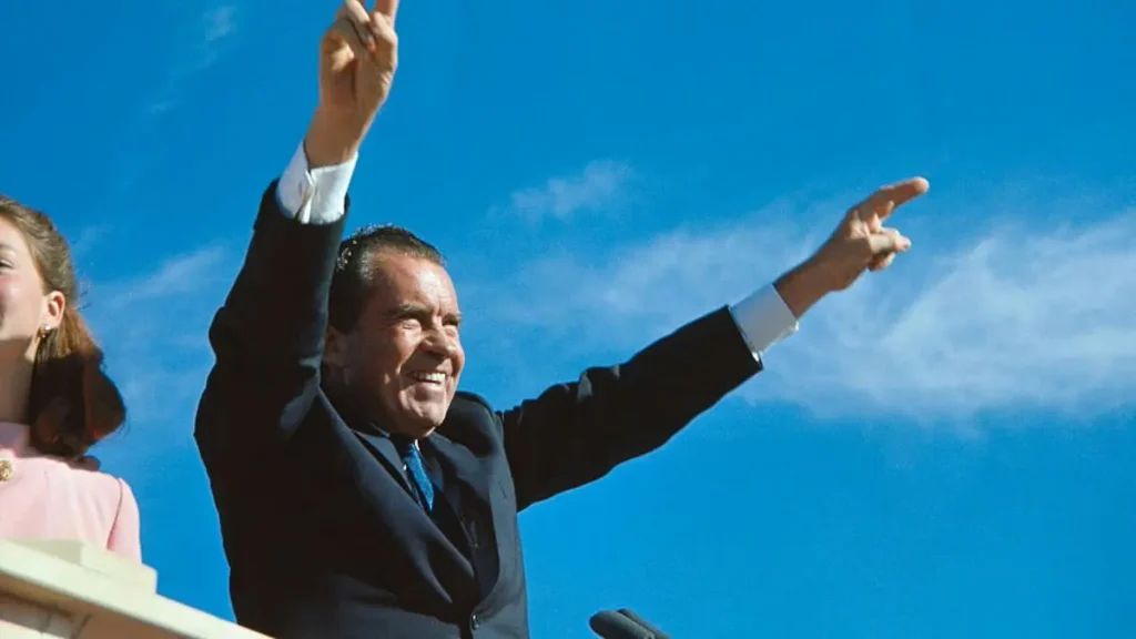 A man in a suit raises his hands in a celebratory gesture against a clear blue sky.