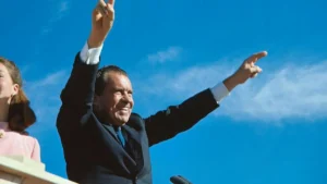 A man in a suit raises his hands in a celebratory gesture against a clear blue sky.