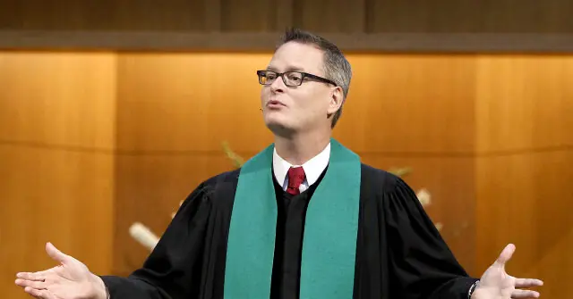 The image shows a person dressed in a black robe with a green stole, gesturing with their hands as if speaking or presenting.