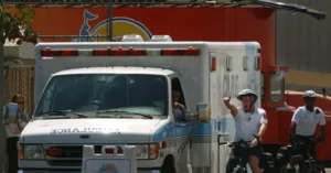 An ambulance is parked near a building with a sign, while two individuals on bicycles gesture towards it.