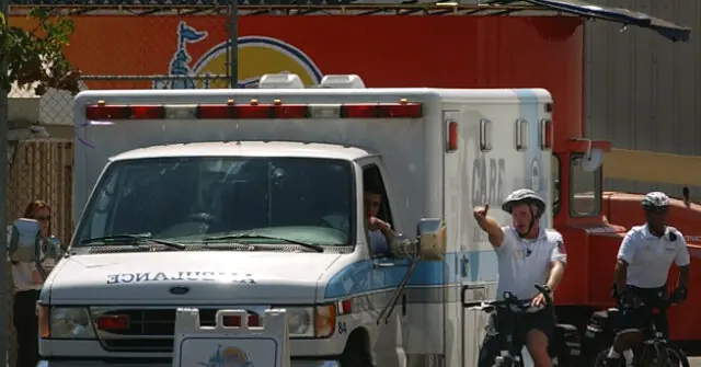 An ambulance is parked near a building with a sign, while two individuals on bicycles gesture towards it.