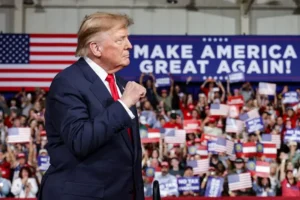 The image shows a political rally with a crowd holding signs and an American flag, with a prominent banner reading "MAKE AMERICA GREAT AGAIN!" in the background.