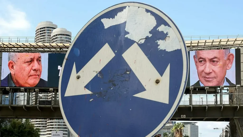 A weathered road sign with two arrows pointing in opposite directions is set against a backdrop of political campaign billboards featuring two prominent figures.