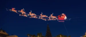 A festive Christmas scene featuring Santa Claus in his sleigh being pulled by reindeer against a dark blue sky.