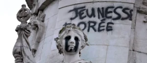 The image shows a vandalized statue with the word "JEUNESSE" spray-painted on the wall behind it.