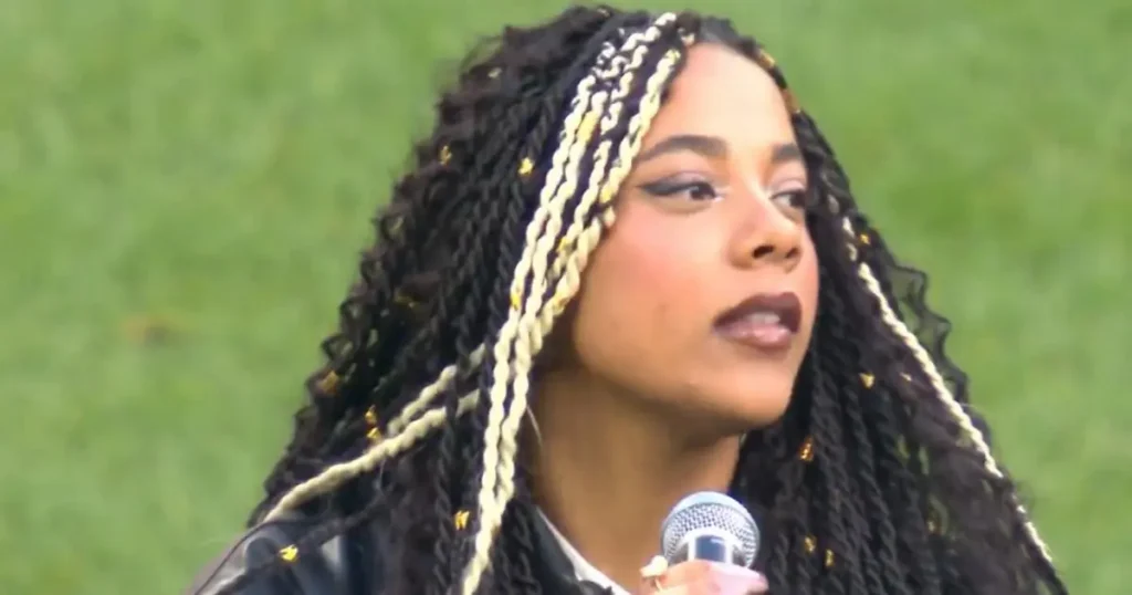 A person with long braided hair is speaking into a microphone, set against a green background.