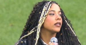 A person with long braided hair is speaking into a microphone, set against a green background.
