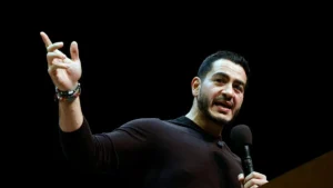 The image shows a person speaking into a microphone, gesturing with their hand, and wearing a black long-sleeve shirt and a bracelet.