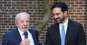 Two men in suits are standing in front of a brick wall, with one holding a microphone and the other smiling.