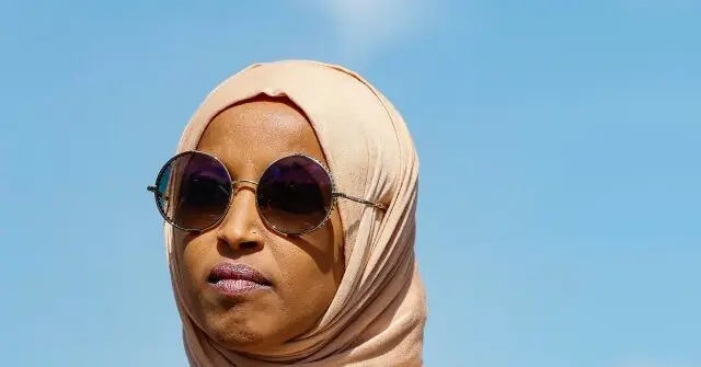 A person wearing a hijab and sunglasses stands against a clear blue sky.