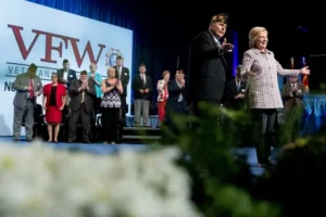 A politician and a veteran are standing on stage at a VFW event, with a group of people clapping in the background.