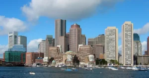 The image showcases a vibrant cityscape with a mix of modern high-rise buildings and historic architecture, set against a backdrop of a clear blue sky with scattered clouds, and a marina filled with boats in the foreground.