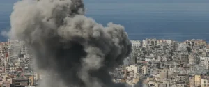 A large plume of smoke rises over a densely packed urban area near the sea, indicating a significant event or incident.