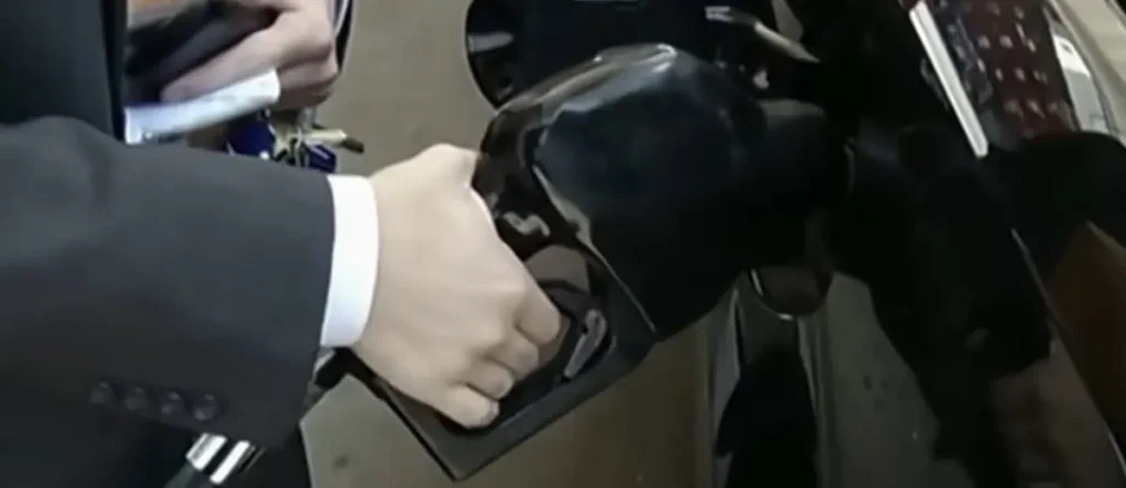 A person in a suit is holding a fuel nozzle at a gas pump.