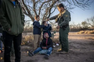 A Border Patrol agent offers water to a child while other individuals observe in a rural setting.