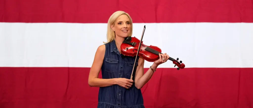 A woman in a denim dress holds a violin in front of a red and white striped backdrop.