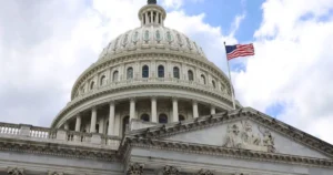The image shows the United States Capitol building with the American flag flying atop it, set against a partly cloudy sky.