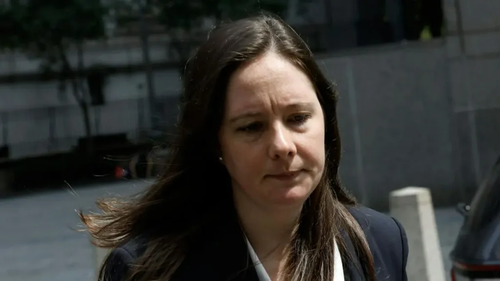The image shows a person with long hair, wearing a dark blazer, walking outdoors on a sunny day.