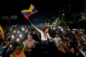 A woman waves a Venezuelan flag amidst a crowd of supporters, illuminated by phone flashlights, during a nighttime event.