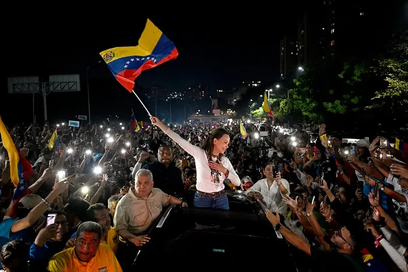 A woman waves a Venezuelan flag amidst a crowd of supporters, illuminated by phone flashlights, during a nighttime event.