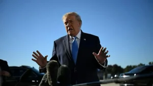 The image shows a man in a suit speaking outdoors, with microphones in front of him, suggesting he is addressing the media.