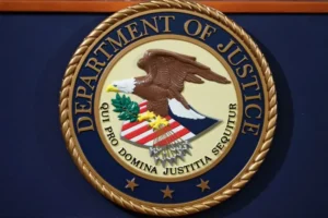 The image shows the seal of the Department of Justice, featuring an eagle, an olive branch, and the Latin phrase "Qui pro domina justitia sequitur."