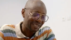 The image shows a man wearing glasses and a striped shirt, smiling and looking to the side.