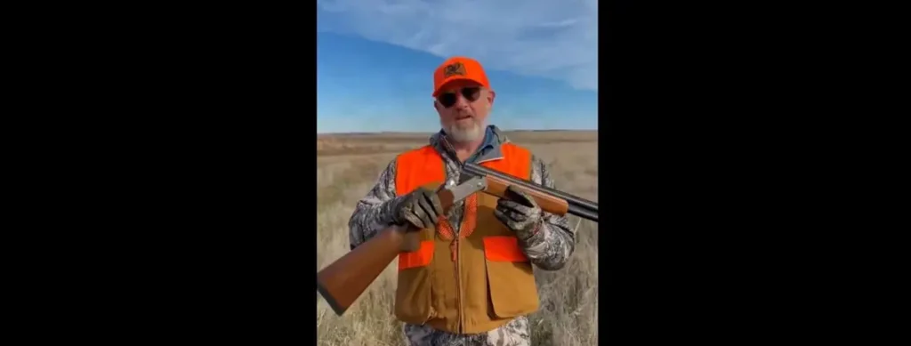 A man in hunting gear holds a shotgun in an open field under a clear sky.