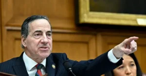 A man in a suit and tie is gesturing with his right hand while speaking at a podium.