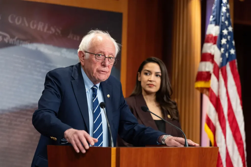 A politician stands at a podium, delivering a speech, with an American flag and another individual in the background.