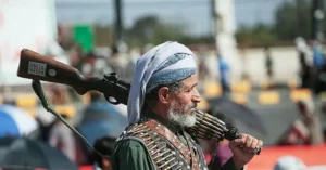 A man in traditional attire carries a rifle and ammunition belt, standing in a public setting with spectators in the background.