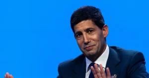 The image shows a man in a suit and tie, gesturing with his hands, set against a blue background.