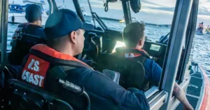 Three U.S. Coast Guard personnel are at the helm of a boat, navigating through a body of water.