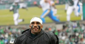 A football player wearing a headband and face paint stands on the sidelines during a game.