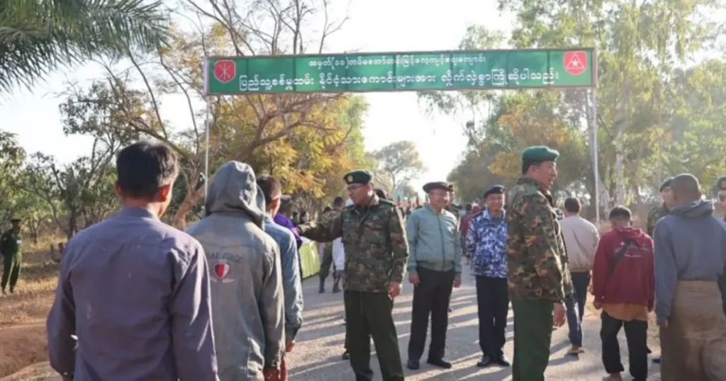 The photo shows a group of people, including military personnel, gathered under a signboard with text in Burmese, indicating a formal or official event.