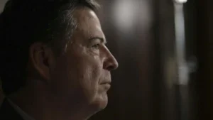 The image shows a close-up of a man in profile, with a serious expression, set against a dimly lit background.