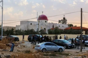 A group of people gather outside a church with a red dome, near parked cars, under a cloudy sky.