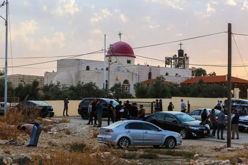 A group of people gather outside a church with a red dome, near parked cars, under a cloudy sky.