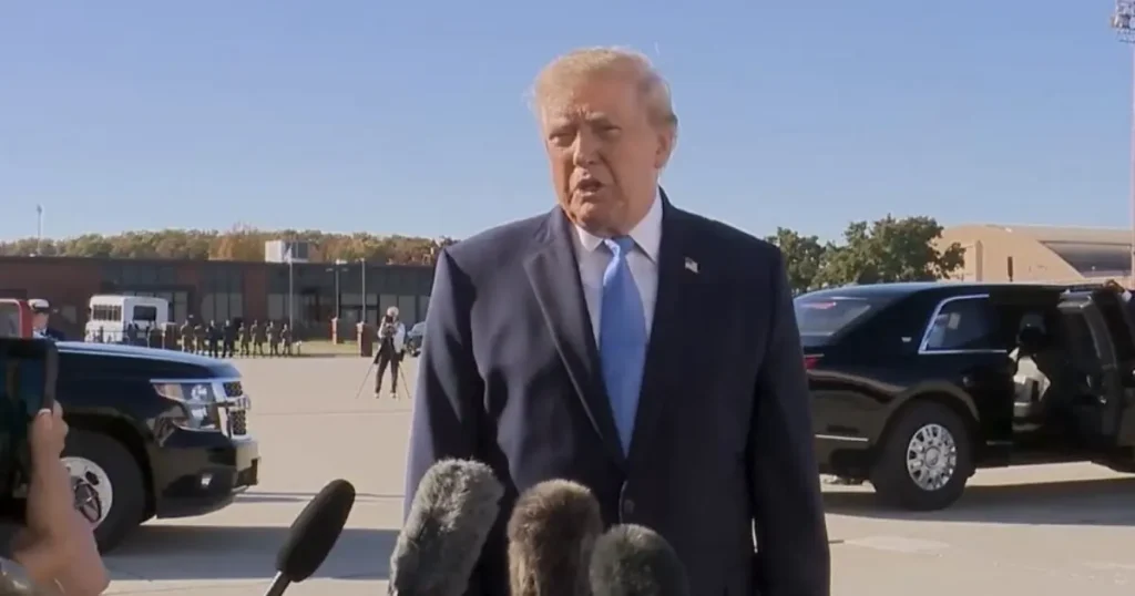 The image shows a man in a suit speaking to reporters in an outdoor setting with vehicles and a crowd in the background.