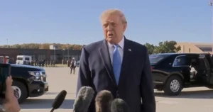 The image shows a man in a suit speaking to reporters in an outdoor setting with vehicles and a crowd in the background.