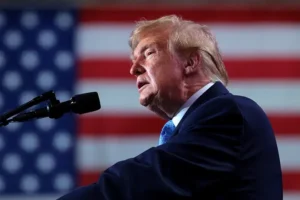 The image shows a man speaking at a podium with an American flag in the background.