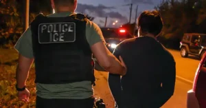 The image depicts a law enforcement officer from ICE (Immigration and Customs Enforcement) apprehending an individual at night.